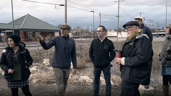 The image shows artist Chris Carl in the middle of a group of four researchers. They are wearing winter jackets and standing on a brown grass field, with the industrial landscape of Granite City in the background. Chris Carl is gesturing with his hands, as he explains his project.