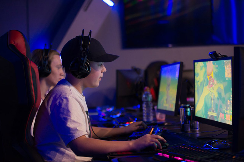 A young man wearing large headphones sits in a gaming chair at a PC playing a game in a dark room filled with purple and blue light.