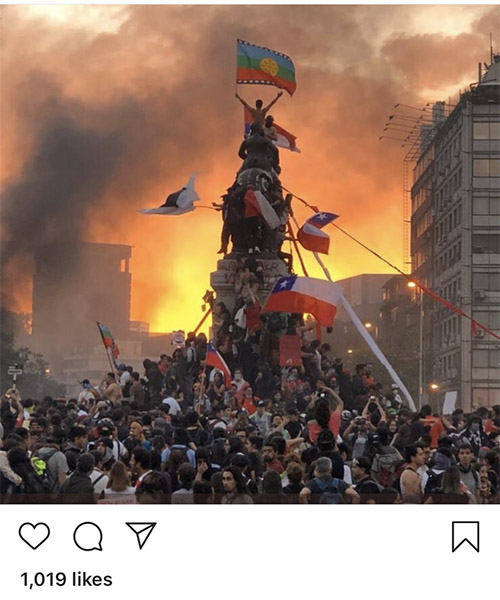 hundreds of protesters surround and climb an obelisk to plant the mapuche flag against and orange sunset