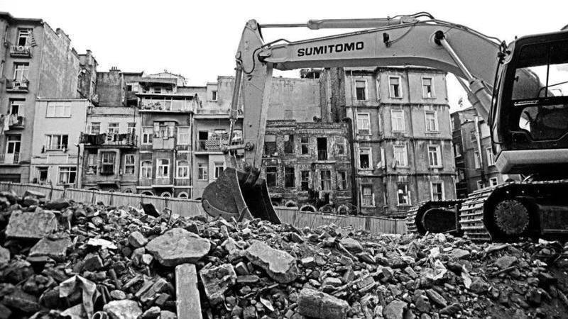 The black-and-white image of a bulldozer with in the midst of the rubble from house demolition. Houses at the background remind of the buildings on the previous image.