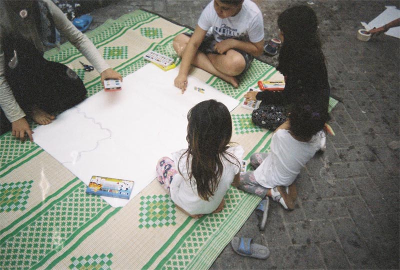 The image shows five children sitting on a rug on a street and drawing with crayons. 