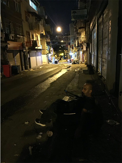 The image of a man sitting in the tire on the right side of a street. It is nighttime, the street is dark. A street light illuminates the street from afar.