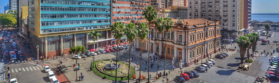 Picture of “Paço da Prefeitura” taken from above. It focuses on the city hall building, built in 1901, the square and the water fountain in front of it. There are tall buildings around it. People are walking on the streets and there are several cars parked. The Guaíba River is spotted on the horizon. 