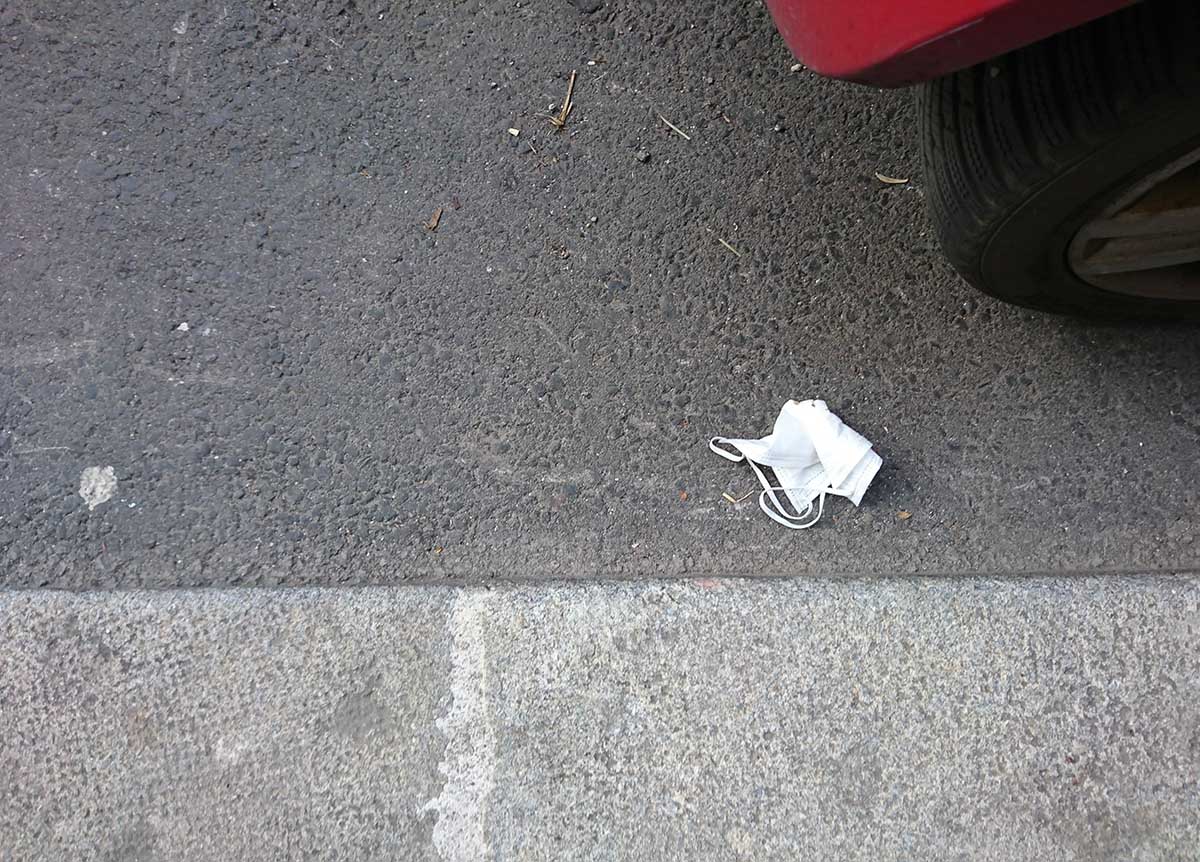 A white face mask lays crumpled on a street. Sticks, rocks, and the cracks of the pavement are visible in this close-up shot. Part of a parked vehicle tire is visible at the edge of the frame.