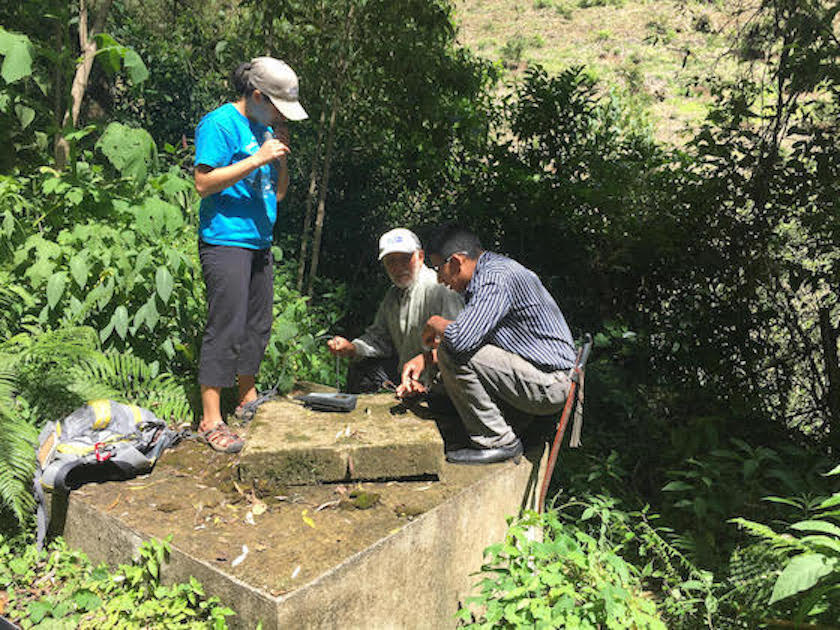 Three people huddled around a cement well in the middle of green foliage, using scientific equipment to measure dissolved oxygen
