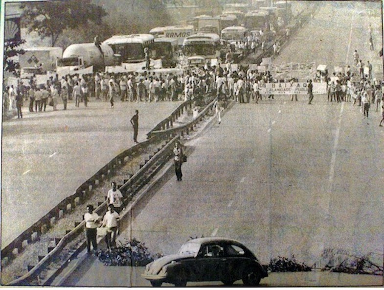 Black and white photo of protestors closing a road holding banners, with a line of vehicles behind them. 