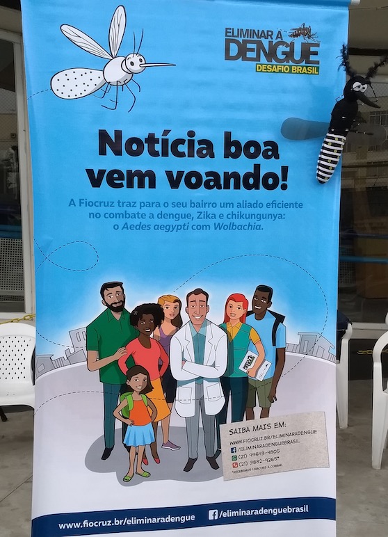 Banner from the WMP project. On the upper right side, it is written “Eliminar a Dengue – Desafio Brasil” (Eliminate Dengue – Brazil’s Challenge), which is the name WMP used in its early days. On the upper left side, there is a white mosquito with green dots. In the middle, in bold, it is written “Notícia boa vem voando” (Good news arrive flying); then, beneath, in smaller font, “A Fiocruz traz para o seu bairro um aliado eficiente no combate a dengue a dengue, Zika e chikungunya: o Aedes aegypti com Wolbachia” (Fiocruz brings to your neighborhood an efficient ally in combating dengue, dengue, Zika and chikungunya: the Aedes aegypti with Wolbachia). Underneath this text there is a drawing of five adults and one child—in the middle is a white man wearing a white coat. On the bottom right, there is a list of places for more information about the technique.