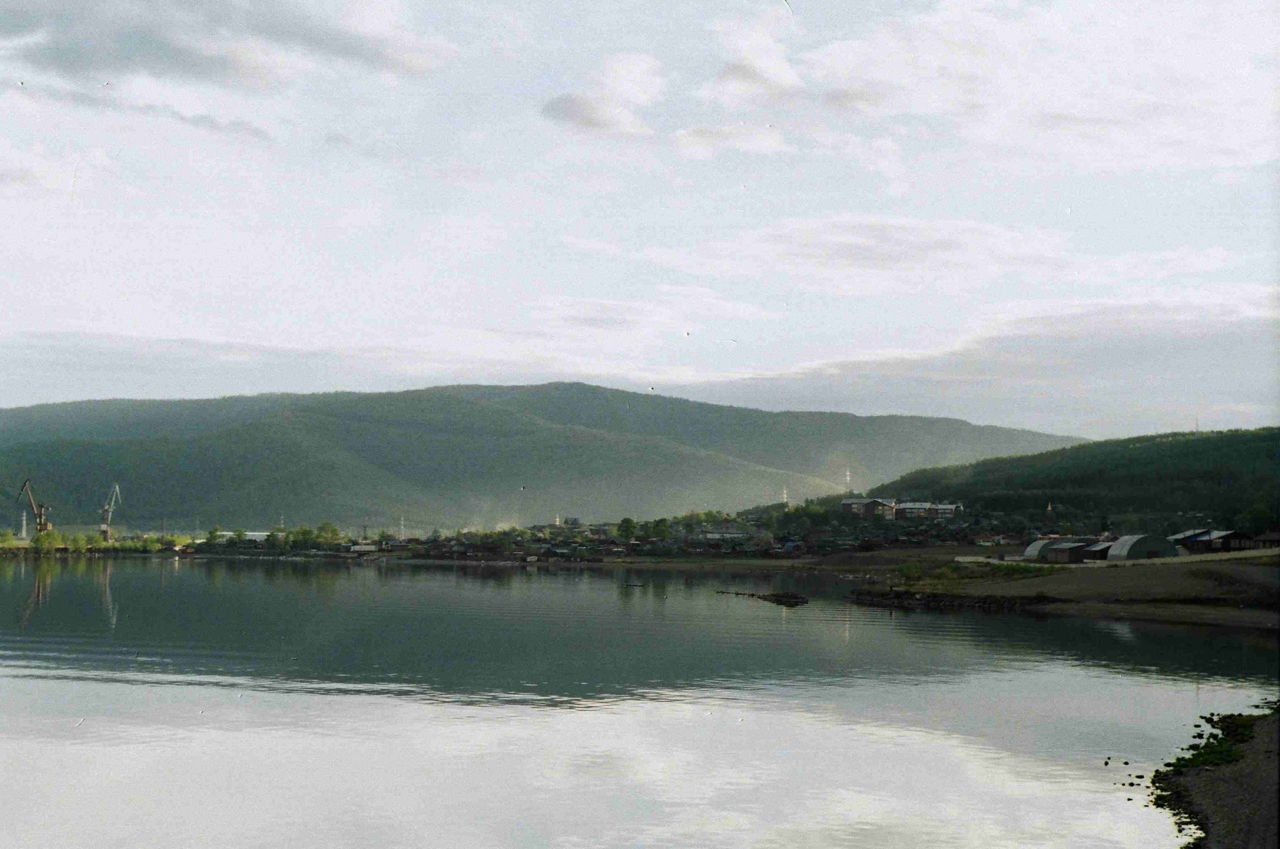 A landscape of the lake shore and the hills behind the lake shore. The water is still and reflects the hills. The sky is cloudy with some patches of blue.