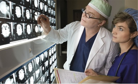 Photo of a male neurologist and a female nurse looking at neurodiagnostic brain scans. The nurse has a stethoscope hanging down from her neck and a folder with documents in her hands. The doctor is pointing at a particular brain image, drawing the nurse's attention to it. 
