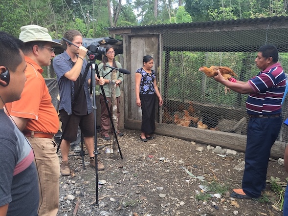 A Maya man is holding a chicken in front of a camera and looks to be explaining something. Behind the camera are a research team and film crew of five people. They are outdoors in a forested area.