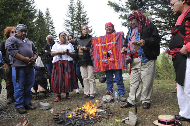Four Maya Elders standing around a small fire. One is holding a colorful red textile that reads, in Spanish, "Great Confederation Ixim Ulew 2020." In the background are other research team members. 