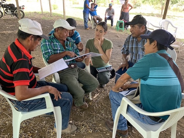 A group of people are sitting in plastic chairs over a flat dirt ground. Some have papers in their hand, they look to be discussing the project.