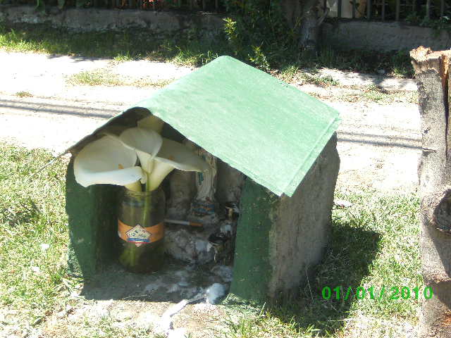 Una animita callejera con forma de casita, con techo verde, flores en un jarrón, y un cigarro.