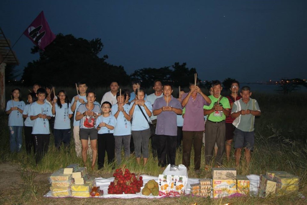 A group of several dozen people, half of them appearing to be students, all holding incense, with offerings of fruit and snacks placed on the ground before them.