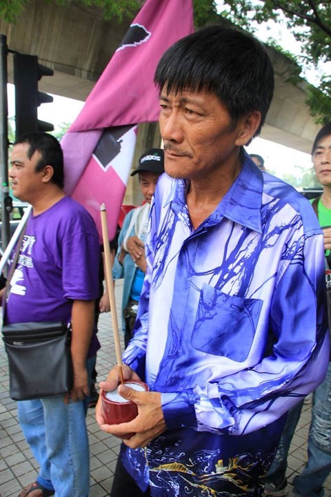 A man wearing a bright purple jacket faces the camera with a solemn look, while holding an incense stick, representing the ancestors.
