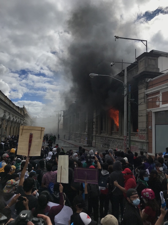 The photo looks over protestors in the street who are standing feet away from the Guatemala Congressional building. The building is on fire, and a smoke plume drifts off into the sky. 