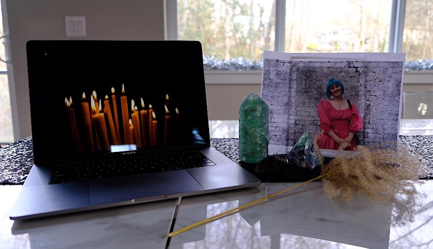 A photograph that shows a laptop and an altar on a counter. The laptop screen shows an image of burning candles. The altar includes a printed photo of the author who is a white woman with blue hair and in a pink dress standing in front of a brick wall. It also includes three crystals and a dried plant. 