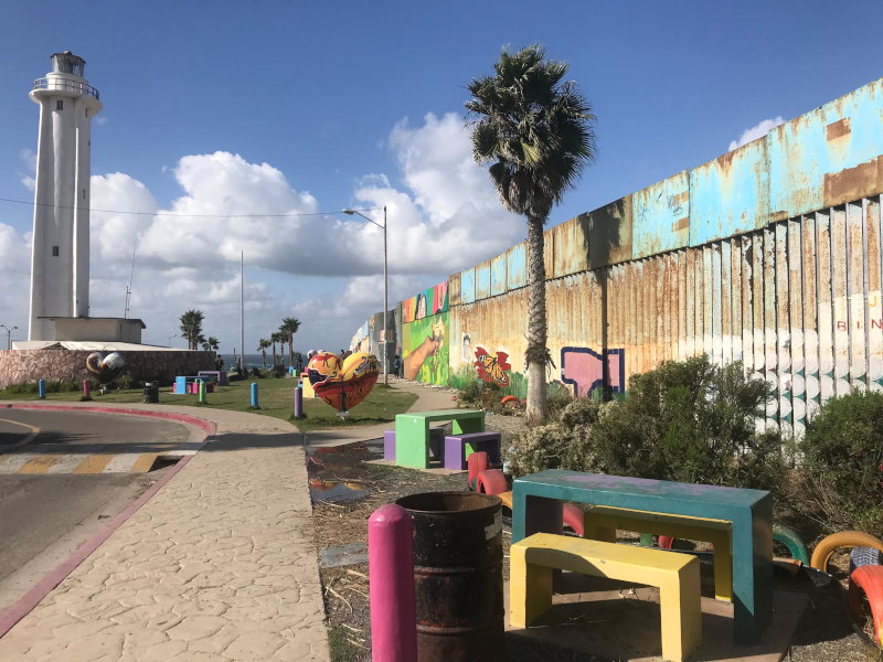 Parque con bancos multicolores y cerca fronteriza y jardín binacional a la derecha. El muro fronterizo tiene un mural brillante de una mano extendida en la distancia.