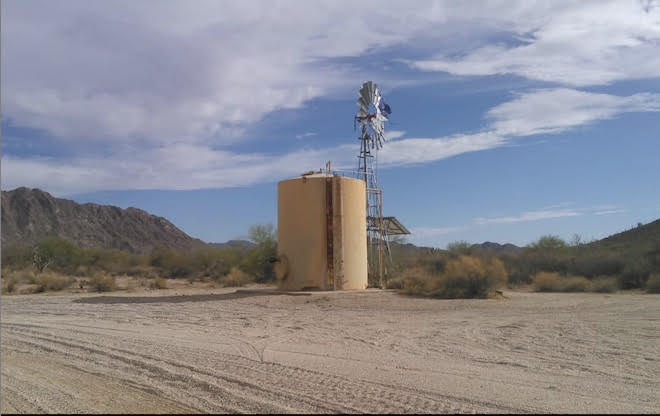 An Image of Papago Well, a large water tank with a faucet. Behind it there is a metal windmill, and barely visible is the purple humane borders flag marking this as a location containing potable water. The water tank sits in a desert landscape, near a sandy road marked by tire tracks.