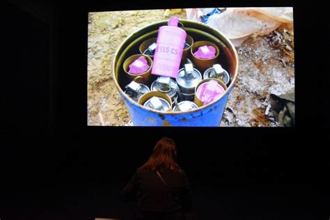 A woman with shoulder length hair stands facing a wall projection of pink tear gas canisters in a blue bucket.