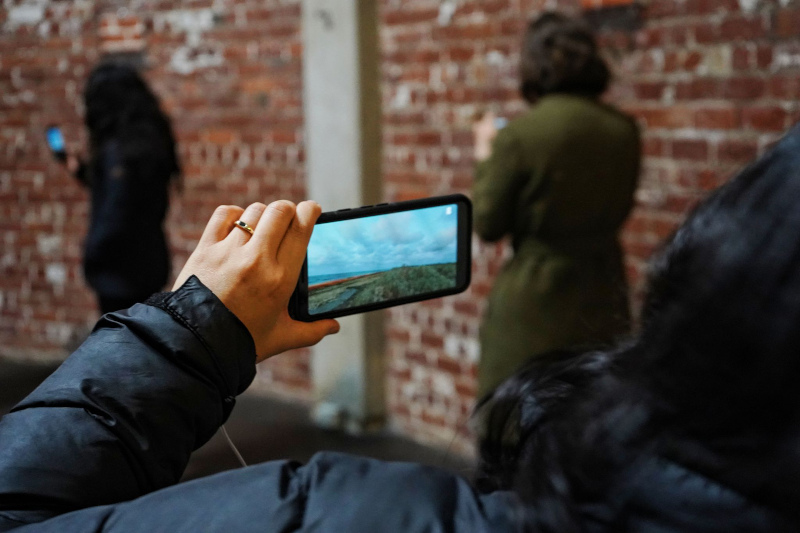 A woman points a smartphone at a brick wall and two other female figures. The screen of the phone shows a landscape with cloudy skies, water, and a green plain. 