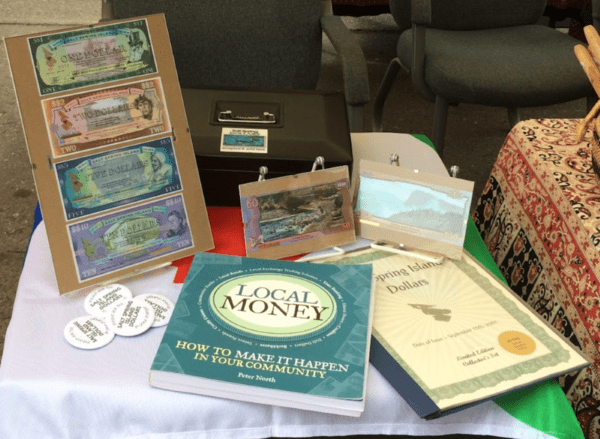 Laid out on a table are copies of the Salt Spring Dollar used within the community of British Columbia along with texts utilized by the author to aid her in gathering information regarding the SSD when doing her research.