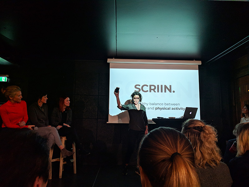 Three women panelists are seated in tall chairs. A woman stands in front of a projector screen holding up a phone. The screen says “SCRIIN”.