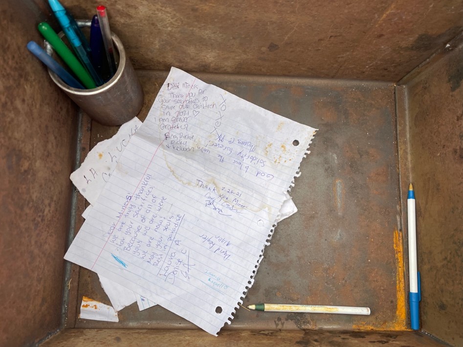 Few pieces of paper with notes to miners on them on the bottom of a mail box at Ludlow Memorial, pencils in a metal glass to the left of the papers, two loose pencils on the right side of the papers