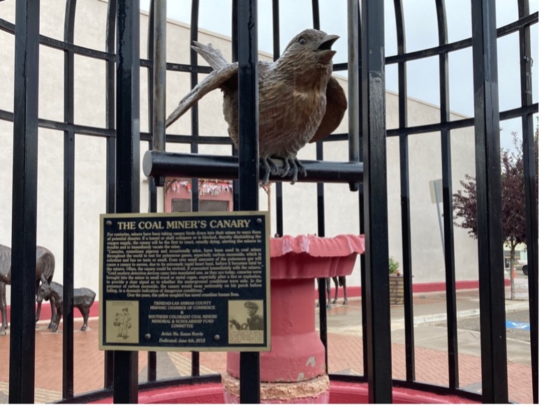 A metal statue of a canary in a cage with a label that reads "The Coal Miner's Canary1