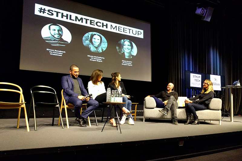 Two co-hosts and three panelists sit in on stage. Behind them is a projection screen that says "#STHLMTECH Meetup" and has photographs, the names, and titles of the three panelists.