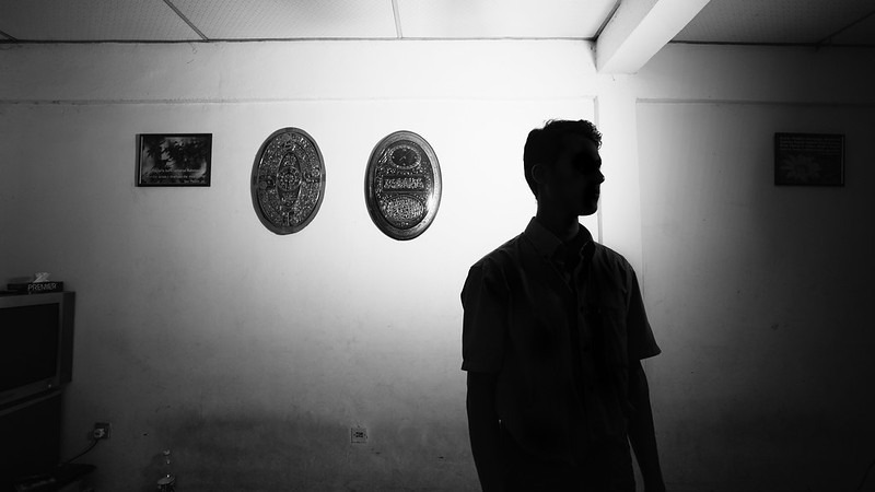 Image consists of a silhouette of a young man who is facing the camera. Behind him are a small series of decorative plates on the wall. The image is in black and white, and the profile of the man is not sharply visible. The man appears to be staring at something slightly away from the camera.