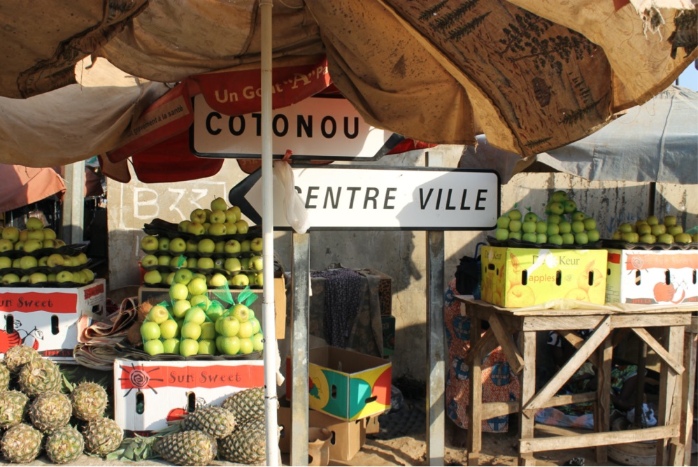 Fruit stand with apples and pineapples. At the back of the fruit stand there are directional signs that point to Cotonou (to the right) and to Centre Ville (to the left) 