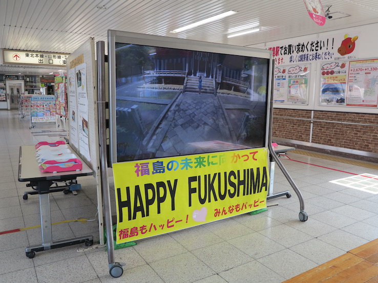 Желтая табличка с надписью Happy Fukushima на английском и японском языках лежит на земле на железнодорожной станции.