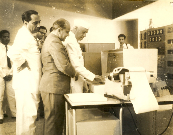 Image of several men in formal attire looking toward India's first digital computer. The image is in black and white. India's first Prime Minister, Jawaharlal Nehru, reaches out to interact with the computer, while four other men close to him look on at the Tata Institute of Fundamental Research. Among the onlookers are Professor M. S. Narasimhan and Homi Bhabha. Away from the computer, against the wall, two other men look toward the demonstration, standing by the edges of the room.