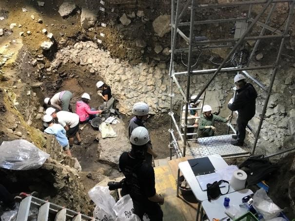 An archaeological excavation site in dark lit by lights, the earth is dug about 3 meters deep, and the site's surface is as big as a standard living room, scattered around 10 archaeologists working together, stairs going down to the site's bottom and other constructions related to the excavation
