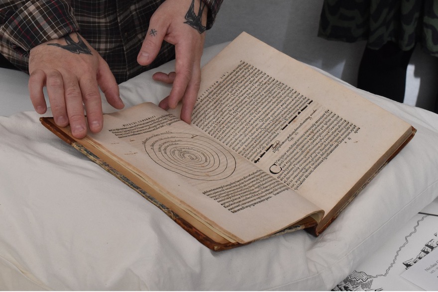 A browned, old book lays open on white cloth. Text and geometric diagrams are visible. Tattooed, light-skinned hands careful reach to turn a page. 