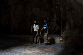Two people wearing backpacks and holding walking sticks stand inside a cave in a dim light.