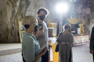 One person stands speaking with gestures in a cave that is brightly lit by a spotlight, while another person looks on recording the conversation with their phone, and a third person wearing a facemask looks directly upwards towards the cave roof.