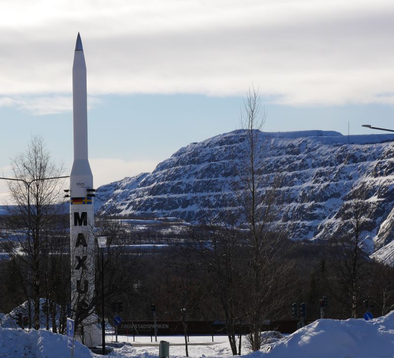 A thin standing rocket with Maxus written from upside down on its bottom half, it is located in the old city center of Kiruna. Around it there are trees without leaves. in the background there is a hill with snow on it, that is where the iron ore mine is located.
