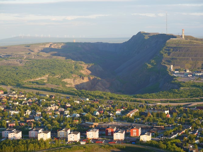 A photo of Kiruna settlement and the ore mine next to the houses on the hill. the photo is taken from far away so that it covers the city, the hill. the city is surrounded by a short green vegetation.further away in the horizon some fading white colored poles that look like windmills.