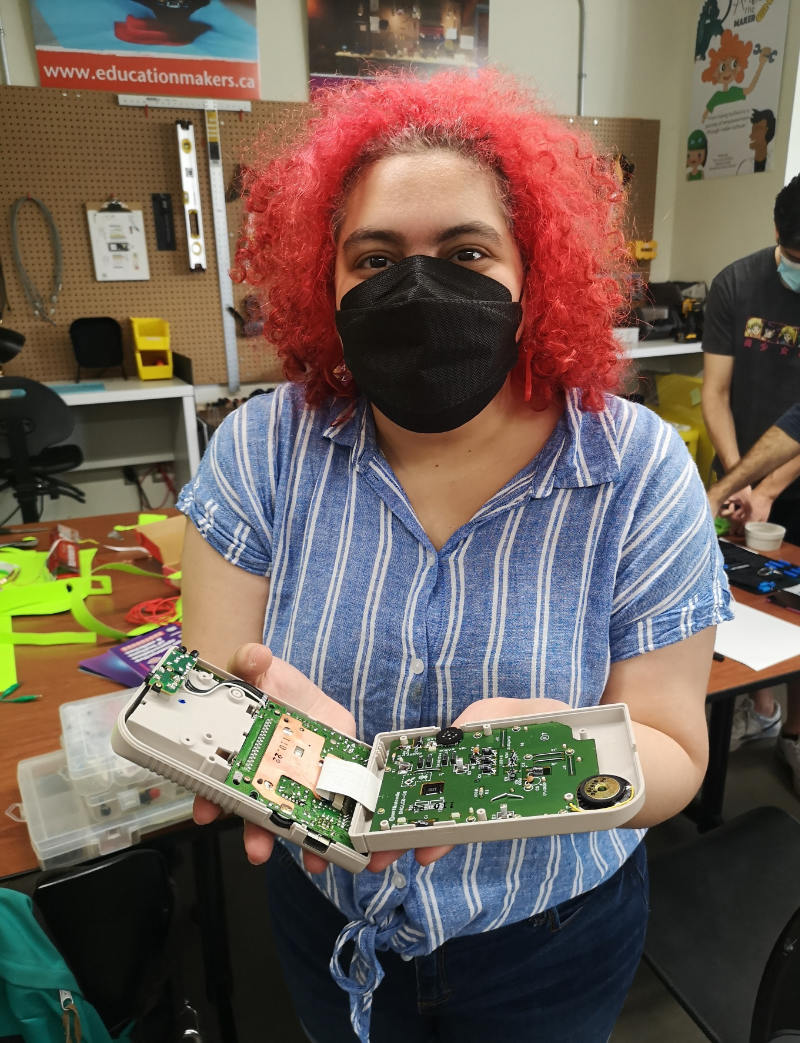 A photograph of a person with dyed bright red hair and wearing a black mask (and a striped shirt). The person has their hands open, and are holding a disassembled games module. Behind them are other kinds of tools for and parts of gaming consoles.