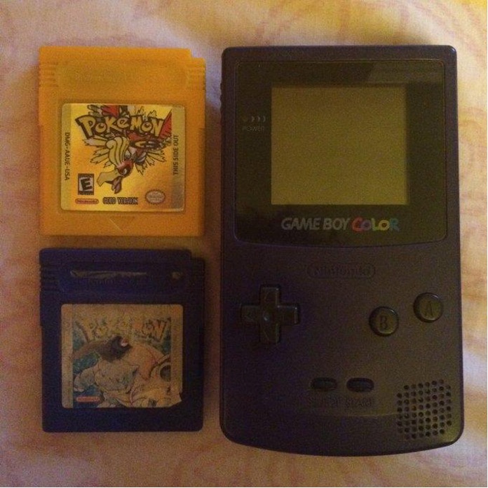 Photo by of a purple Game Boy Color with Pokémon Gold (in yellow) and Pokémon Blue (in blue) cartridges. The photo is on a fabric background.