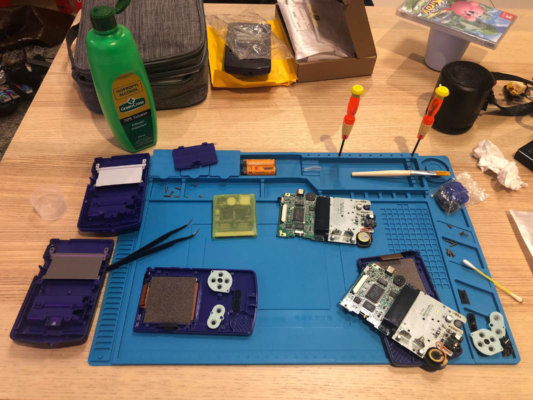 A photograph of a disassembled Game Boy on a bright blue pad, on a wooden desk. There are various tools and parts in the frame.