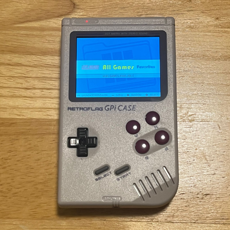 A photograph of a classic Game Boy-style emulator case, with grey case and buttons. The screen is glowing and says "All Games."