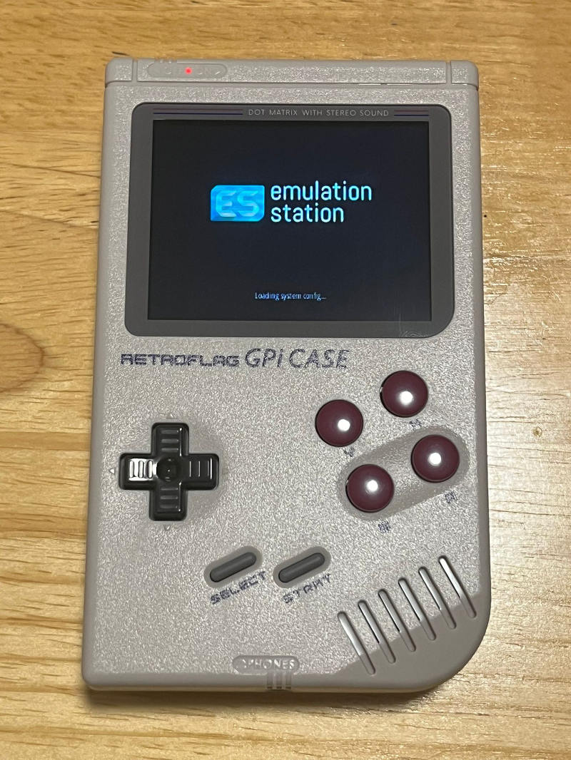 A photograph of a classic Game Boy-style emulator case, with grey case and buttons. The screen is glowing and says "Emulation Station."