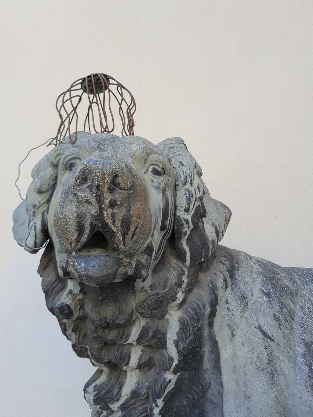 A statue of a proud-looking gray dog with white and brown rivulets of discoloration from age. A wire cage sits upside down on its head. 