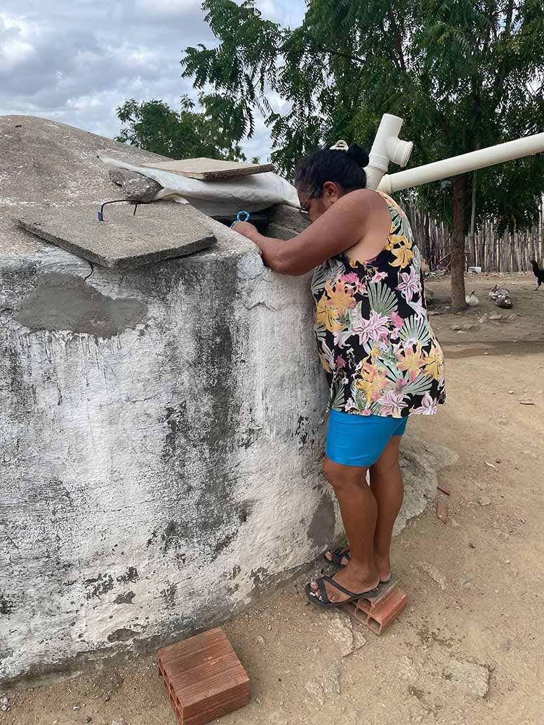 A short woman in a flower patterned sleeve-less shirt and blue shorts fetches water from the opening of a gray/white cylindrical cistern using a barely visible blue rope. The cement cover for the opening is resting on the cone shaped top of the cistern. 