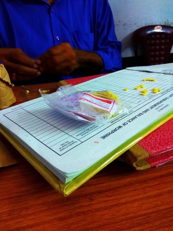 Man in blue shirt sorting out morphine tablets present from a small polythene bag placed over a ledger to record the number of tablets disbursed. 