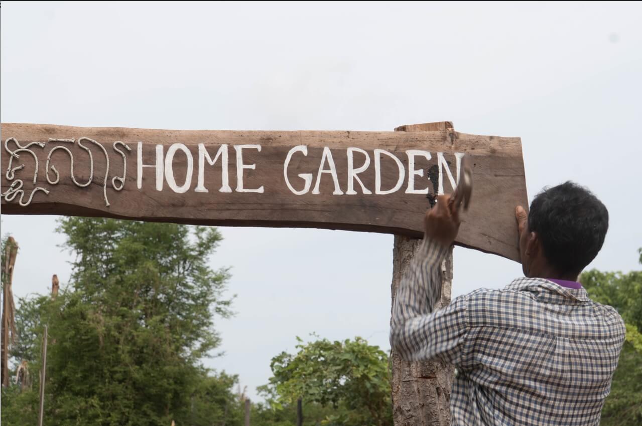 A sign reading "Home garden" in English and Khmer is being adjusted by a man with his back turned towards the viewer.