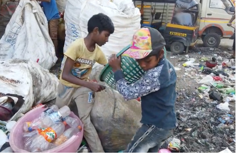 Two young boys sorting through waste amidst large bags of recyclables. The scene includes scattered trash, a pink basin with plastic bottles, and a vehicle in the background.